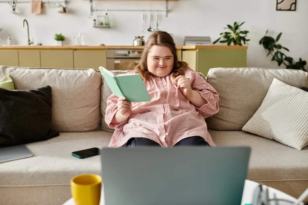 Disabled woman with down syndrome sitting on couch at home using notebook and laptop